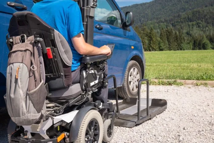 Invalid na električnem invalidskem vozičku pelje proti vrtljivemu dvigalu, ki ga dvigne in zavrti v kombi vozilo.