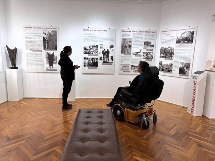 Kustosinja in moški na invalidskem vozičku si ogledujeta kronologijo izdelave spomenika v Zasavskem muzeju Trbovlje.